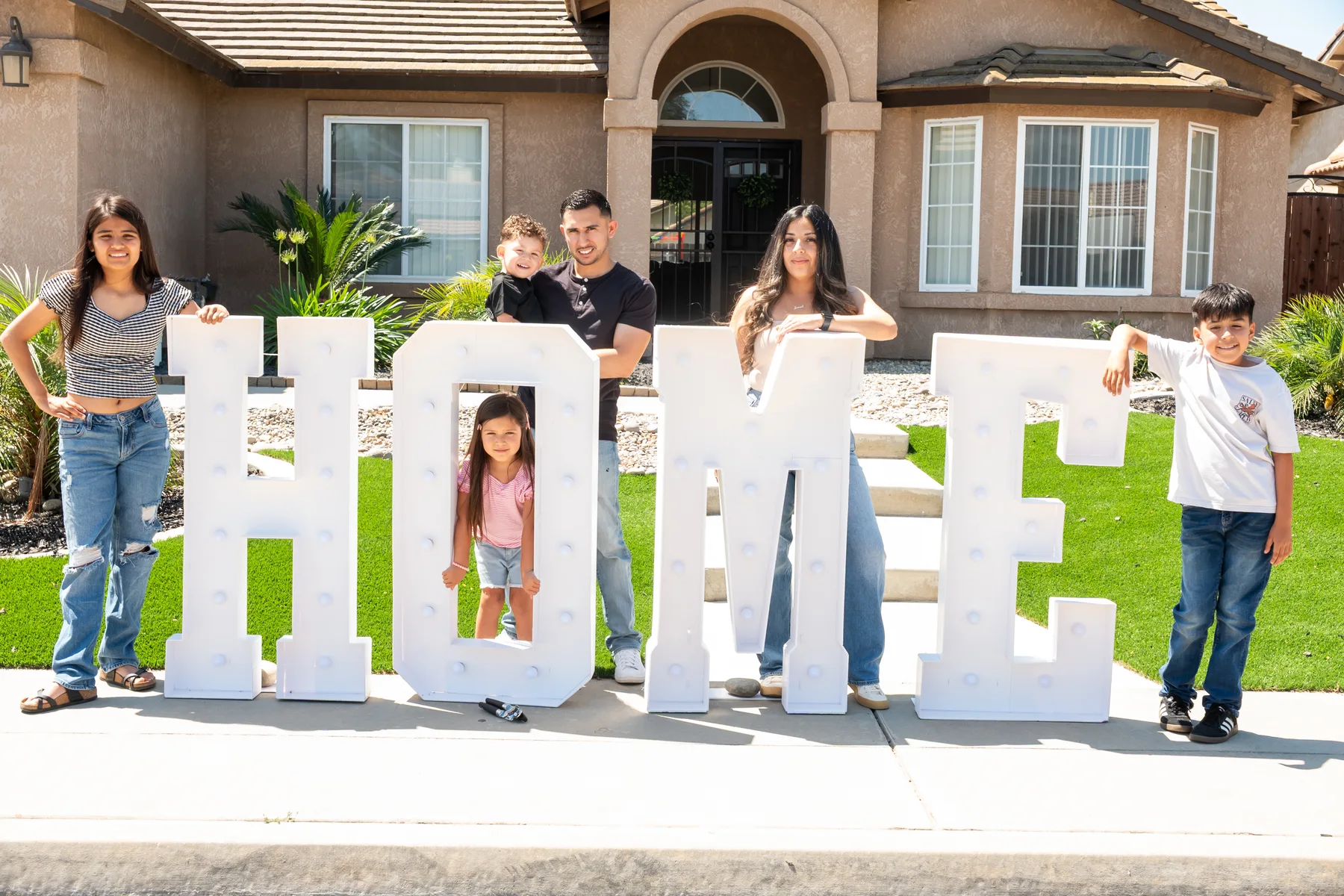 Group stands together around a HOME sign on the lawn.